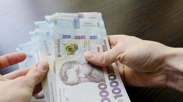 Close up of hands counting Ukrainian hryvnia banknotes on dark wooden table. 