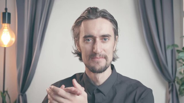 Applauding man with mustache smiles and gently claps while performing slow hand movements against softly lit background with gray curtains, filament bulb, and small plant maintaining warm eye contact