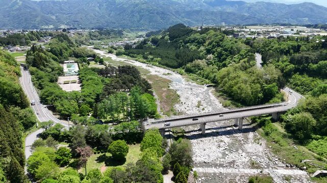 Aerial view of a concrete bridge spans a dry riverbed. Lush green hills surround the scene. Roads curve gently through the landscape. Mountain range rises in the background