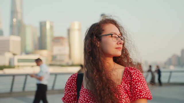 Young woman riverfront skyline sunset contemplative, curly hair and glasses in red dress, city skyscrapers blurred in background, pedestrians passing on promenade, warm golden light, slow motion