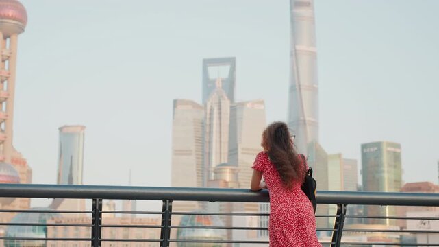 Woman on waterfront railing studying tower skyline at dusk, red patterned dress, city towers and oriental pearl visible, framing shot and soaking in evening glow and reflective glass facades