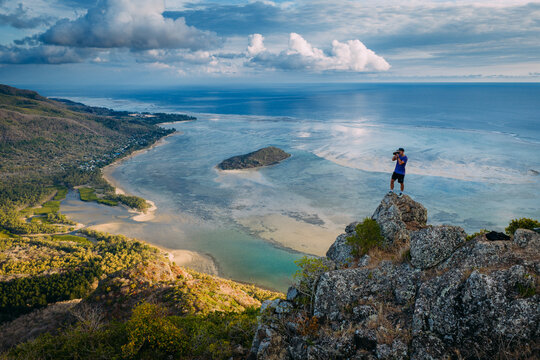 Aerial view of a photographer standing on a rocky mountain peak overlooking the turquoise lagoon and lush tropical coastline in Moka District, Mauritius.