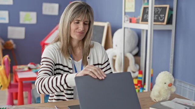 Blonde middle age woman teacher closes laptop, places hands behind head and stretches at a desk in a preschool building filled with toys and a chalkboard; relief pause.