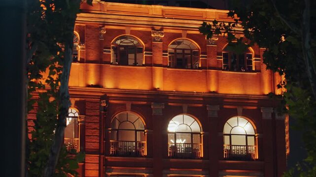Historic brick facade glowing in amber light, arched windows revealing warm interior silhouettes, ornate cornices and balconies framed by street trees, vintage lanterns casting patterned shadows,