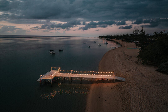 Aerial view of a wooden pier decorated with glowing string lights on a sandy beach at dusk with small boats anchored in the calm bay in Mauritius.