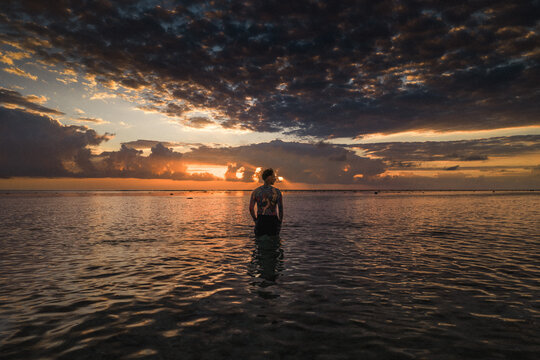 Aerial view of a man with tattoos standing in the shallow ocean water during a dramatic sunset with dark clouds over the horizon in Moka District, Mauritius.