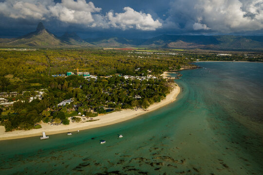 Aerial view of a pristine sandy beach and turquoise lagoon bordered by lush tropical vegetation and dramatic mountain peaks under a cloudy sky Moka District, Mauritius.