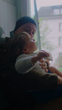 Mother undergoing chemotherapy holding toddler by window at home, headscarf parent supporting child on lap, caregiving moment, maternal bonding, resilience and quiet family connection