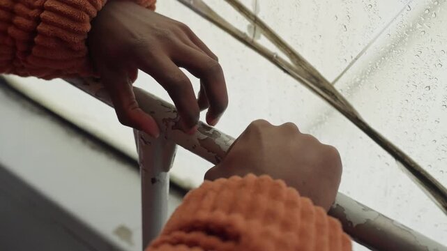 Black hands opening skylight in rain, closeup of orange sweater gripping latch and metal frame, raindrops on glass and rooftop drip visible sequence reads like building caretaker securing shelter,