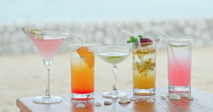 Tropical cocktails with fruit garnishes sitting on wooden table, sandy beach and blurry sea background, summer vacation travel promotion