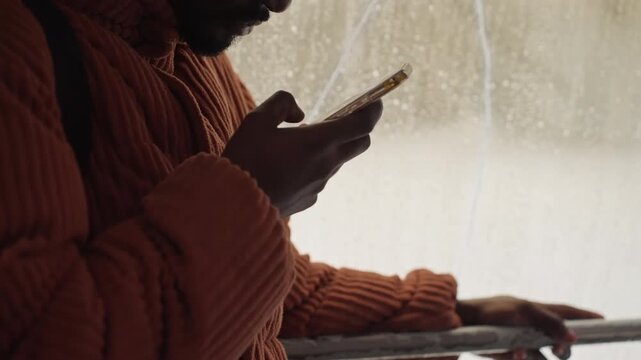 Side profile black man checking phone, beanie and glasses, orange sweater, backpack strap, rainstreaked window, pensive commuter portrait, soft backlight, urban winter mood, solitude and reflection