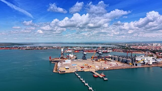 Approaching the modern commercial water-side facility with diverse equipment. Some boats are near the quay in the port. Fluffy clouds in the sky.