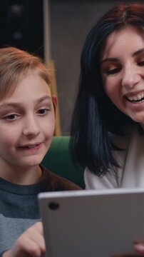 Happy young woman cuddling talking to small kid son, having fun together on couch while using tablet device sitting on sofa in kitchen room. Slow motion