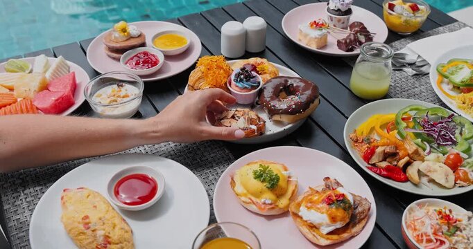 Hand reaching for almond pastry on wooden table full of varied food plates, outdoor resort pool background, overeating and luxury breakfast concept, binge eating lifestyle