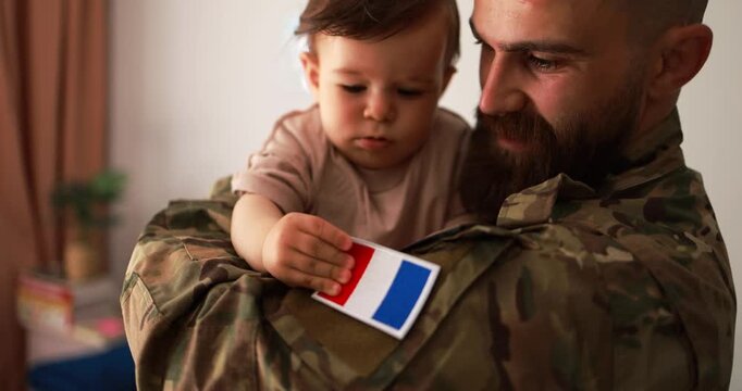 Male french soldier in military uniform hugs his little son at home while baby boy touching french flag - Emotional military happy homecoming concept