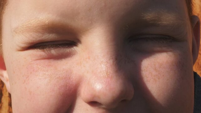 Portrait of little red-haired boy staring and squinting with happy sight. Male face of child illuminated by sun looking into camera and laughing. Facial expression of smiling kid with positive emotion