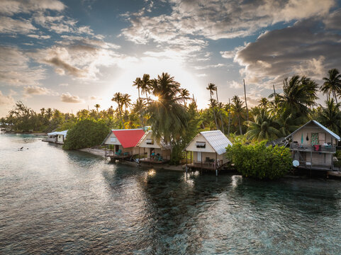 Aerial view of tropical bungalows along the turquoise shoreline surrounded by lush palm trees under a glowing sunset sky Moorea-Maiao, Windward Islands, French Polynesia.