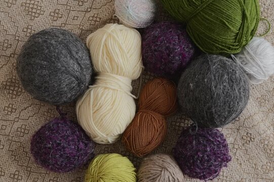 Different balls of yarn and wool are spread out on a light patterned surface. The yarn comes in many colors and textures suited for a variety of crafting tasks