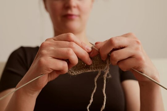 A woman sits comfortably indoors, working on a knitting project. She uses two needles and focuses on her work while yarn hangs down. She enjoys a simple moment at home