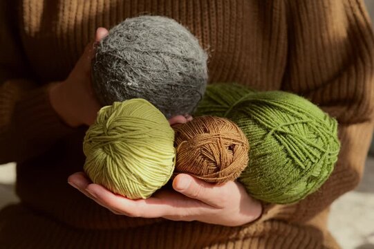 A person with a brown sweater holds four balls of yarn in colors of green, brown, and gray. Sunlight enters the room, creating warm lighting around the yarns