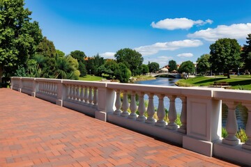 Fototapeta premium White concrete balustrade on brick paved terrace overlooks calm blue river, green park, bridge, and buildings on summer day with fluffy clouds.