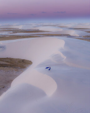 Aerial view of Parque Nacional dos Lencois Maranhenses with a woman in a blue dress standing on white sand dunes under a purple sunset sky in Lencois Maranhenses, MA, Brazil.