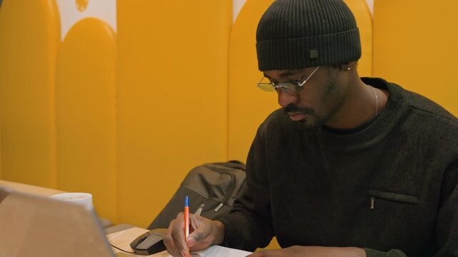 Black designer sketching at laptop desk, creating concept drawings on paper, colored pens and glasses on table, yellow partition background, collaborative studio vibe, freelance brainstorming