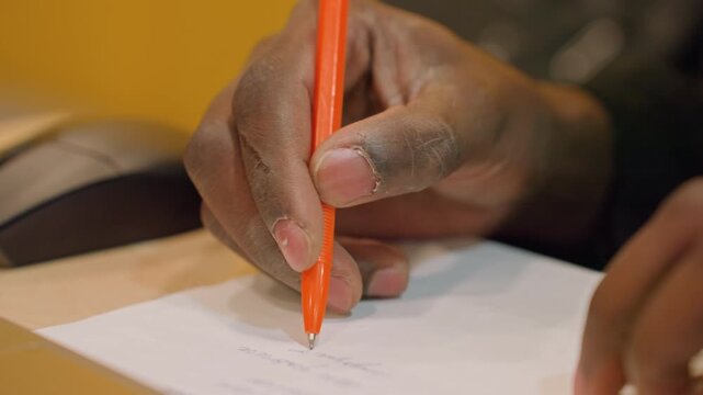 Close inspection of signature on official document. Person signing contract with pen near computer equipment. Detailed view of hands signing legal paper with electronic devices around