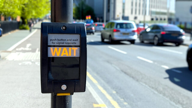 Pedestrian crossing push button close up with illuminated wait sign, UK street traffic control system with blurred cars background