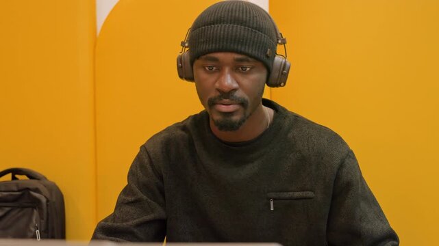 Black man with laptop, yellow booth, engaged audio editor reviewing tracks on screen, wearing beanie and headphones, backpack by seat, warm studio lighting, remote collaboration vibe with focused