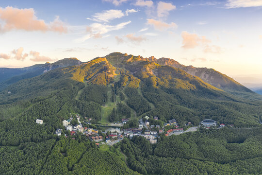 Aerial view of Cerreto Laghi mountain resort nestled in dense green forests and ski slopes under a golden sunset sky, Cerreto Laghi, Emilia-Romagna, Italy.