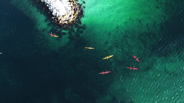 Top-down aerial view of a group kayaking in crystal clear turquoise arctic waters by a rocky pier in Lofoten Islands, Norway. Active outdoor adventure in the scenic Scandinavian fjords