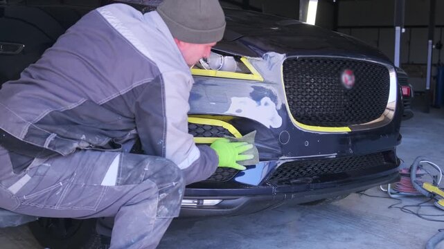 Auto technician inspecting repaired car bumper in workshop after sanding and masking process, preparing damaged surface for primer coating and repainting.
