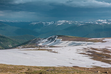 Sunlit landscape with small lake among snowfields, thawed ground, forest and snow cornice on long spur with view to big snowy mountain range far away under cloudy sky. Sunlight and shadows of clouds. © Daniil