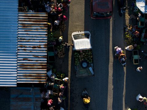 Aerial view of a white pickup truck carrying coconuts through a busy street market with people and stalls in Kemalang, Central Java, Indonesia.