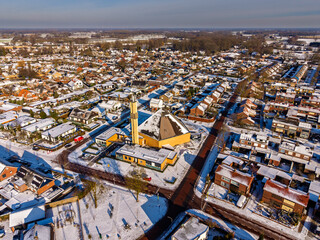 Fototapeta premium Aerial view of a snow-covered Dutch town with a modern church building and residential houses under a clear blue sky. Winter landscape in the Netherlands with snowy rooftops and streets.