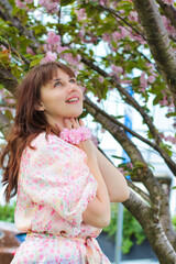 Fototapeta premium Portrait of a brunette woman standing by blooming sakura trees in a park. Medium portrait of a brunette woman in a soft style outfit standing in a sunny park with blooming sakura trees. Natural dayli
