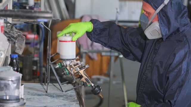 Professional auto body painter tightening paint cup lid on spray gun before automotive primer application during vehicle refinishing and repair process in workshop. Spray gun assembly