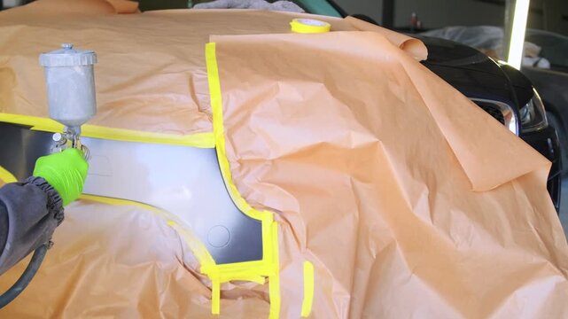 Auto body repair technician spraying primer on masked front bumper during vehicle paint preparation and automotive body repair process in garage workshop. Primer spraying process