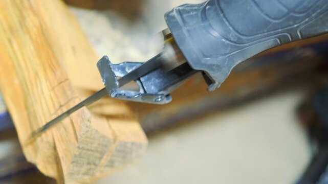 Electric reciprocating saw cutting thick wooden beam during active construction work. Close-up of fast moving blade slicing timber material in workshop environment.