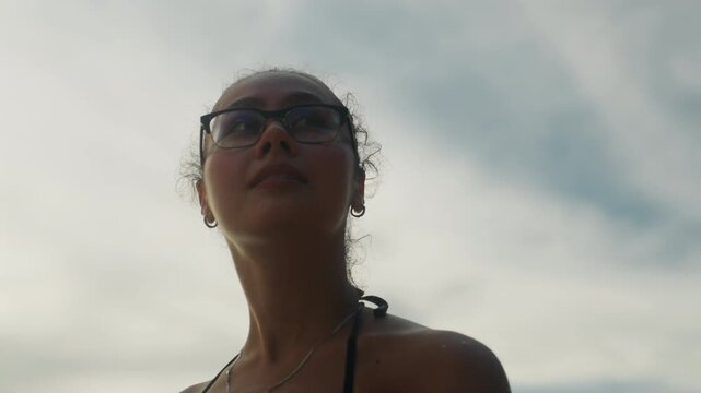 Young woman looking skyward at dawn, reflective solo traveler on beach, soft backlight and clouded horizon, glasses silhouette, calm expression and gentle breeze, minimal emotional tone.