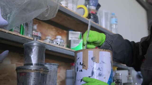 Auto body technician mixing automotive paint with stir stick inside metal container before preparing spray gun equipment for vehicle repainting in workshop.