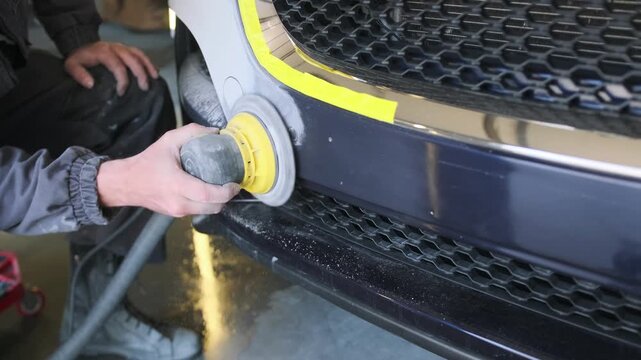 Professional auto body technician polishing repaired front bumper with electric orbital machine before automotive repainting and finishing process in workshop. Car Machine polishing before painting