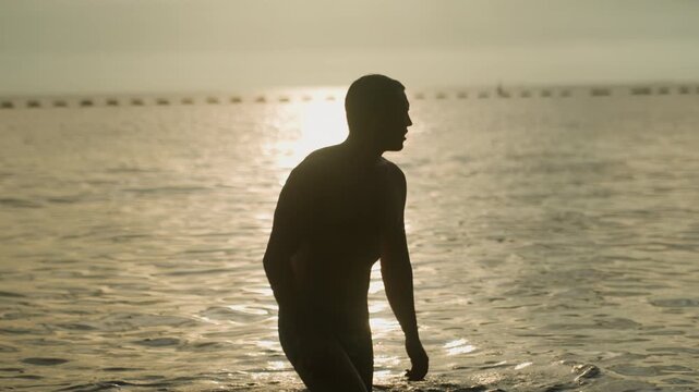 Male silhouette walking out of water, sunset backlight casting long glow, purposeful stride and dripping droplets, strong posture and triumphant mood at shoreline, cinematic athletic energy