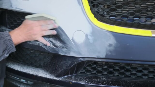 Professional auto body technician hand sanding repaired bumper surface after machine polishing before automotive repainting and finishing process in workshop. Hand finishing