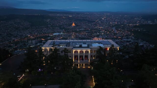 Aerial night view of Mtatsminda Park restaurant and funicular station in Tbilisi, Georgia, featuring illuminated arched architecture