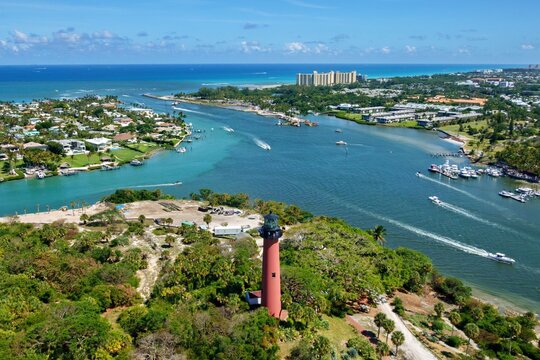 Aerial view of the Jupiter Inlet Lighthouse overlooking the turquoise waters of the inlet with boats cruising past lush coastal greenery in Jupiter, Florida, United States.
