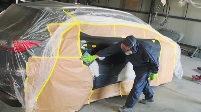 Professional auto body technician wiping repaired vehicle door before automotive painting and refinishing process in collision repair workshop with masked car body. wiping repaired car door 