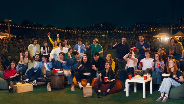 Young people cheering at outdoor night watch party with drinks and snacks. Concept of live sports viewing, fan celebration, summer festival, social gathering and entertainment ads.