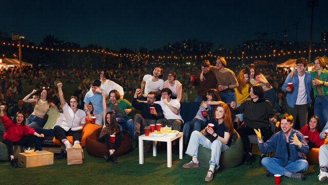 Excited crowd of friends celebrating outdoor sports viewing party at night. Concept of fan zone, live match broadcast, youth gathering, summer night leisure and event promotion.